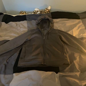 Grå fleecejacka från Sail Racing - Säljer en grå fleecejacka från Sail Racing med dragkedja och huva. Hoodie har en bröstficka med dragkedja och är perfekt för kyliga dagar. Den är både stilren och funktionell.