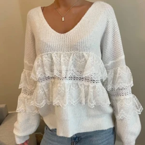 Vit stickad tröja med spetsdetaljer - Säljer en stilren vit stickad tröja med v-ringning och vackra spetsdetaljer på ärmarna. Tröjan har långa ärmar och volanger. 