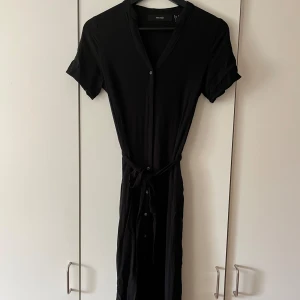 Svart klänning med knappar och knytband - Säljer en stilren svart klänning med korta ärmar och v-ringning från vero moda. Klänningen har knappar framtill och ett knytband i midjan för en snygg passform. Perfekt för både vardag och fest!