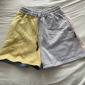 Flerfärgade shorts från Playboy - Säljer ett par unika flerfärgade shorts från Playboy. De har en cool design med olika färger på varje ben: gul, ljusgrå, rosa och blå. Shortsen har en elastisk midja med snörning för bästa passform. Perfekta för en avslappnad stil!
