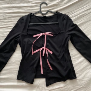 Svart topp med rosa rosetter - Säljer en snygg svart topp med långa ärmar och två rosa rosetter framtill. Toppen har en stilren design med en liten öppning nedtill.