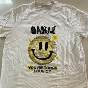Vit t-shirt med smiley från Ganni - Säljer en cool vit t-shirt från Ganni med ett stort smiley-tryck i gult och svart. Texten 'You're gonna love it' står under smileyn. Perfekt för en avslappnad stil och passar till alla tillfällen!