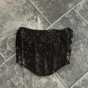 Svart glittrig korsett - Snygg svart korsett med glittriga paljetter och snörning i ryggen. Perfekt för festliga tillfällen när du vill sticka ut. Den har en tight passform och är axelbandslös för en elegant look. Storlek M men passar även mindre och större eftersom man justerar själv.