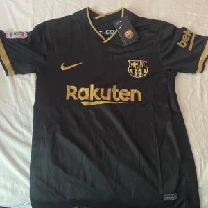 Svart och guld FC Barcelona tröja - Säljer en snygg FC Barcelona tröja i svart med guldiga detaljer. Tröjan har korta ärmar och är i nyskick. Den har klubbens emblem och sponsorlogga i guld på framsidan. Perfekt för alla Barca-fans!