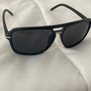 Svarta solglasögon med metallbrygga - Helt nytt  Använt 1 gång   Snygga svarta solglasögon med en cool metallbrygga över näsan. De har en klassisk rektangulär form och ger en stilren look. Perfekta för soliga dagar och passar till alla outfits!