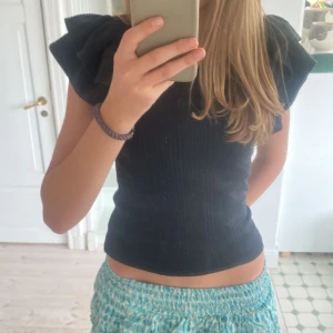 Svart ribbad topp från Zara - Säljer en superfin svart ribbad topp från Zara. Den har korta ärmar med volangdetaljer som ger en extra touch. Perfekt för både vardag och fest! Toppen är i storlek S och sitter tight.