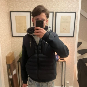 Mörkblå jacka från Sail Racing - Säljer en mörkblå jacka från Sail Racing med huva och dragkedja. Jackan har ett broderat märke på bröstet och är tillverkad i syntetmaterial. Perfekt för kyligare dagar.