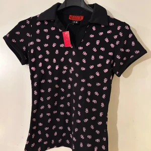 Svart och rosa pikétröja från Killstar - Snygg svart pikétröja från Killstar med rosa dödskallemönster. Tröjan har korta ärmar och en klassisk krage. Perfekt för dig som gillar en edgy stil. 🖤