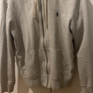 Grå hoodie från Ralph Lauren - Säljer en stilren grå hoodie från Ralph Lauren med dragkedja och klassisk logga på bröstet. Tröjan har långa ärmar och en praktisk huva med snören. Perfekt för en avslappnad look. Väldigt populär kofta ute på marknaden säljs inte längre på de flesta butikerna väldigt sällsynt. 