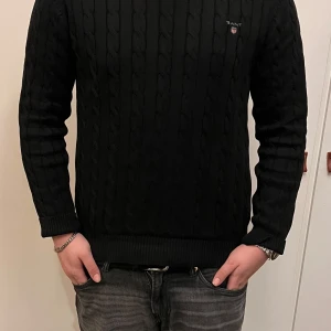 Svart kabelstickad tröja från GANT - Säljer en stilren svart kabelstickad tröja från GANT. Tröjan har en klassisk design med GANT-loggan broderad på bröstet. Perfekt för kyligare dagar med sin varma och bekväma passform.