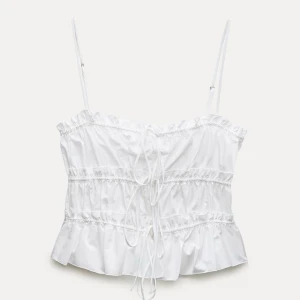 Vit croppad topp med volangdetaljer - Säljer en söt vit croppad topp med smala axelband och volangdetaljer. Toppen har snörning framtill och en luftig passform, perfekt för varma dagar.