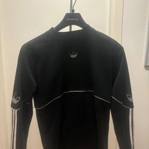Svart långärmad tröja från Adidas - Säljer en svart långärmad tröja från Adidas med vita detaljer och logga på bröstet och ärmarna. Tröjan har de klassiska tre ränderna längs ärmarna och en rund halsringning. 8/10 kond- passar xs och S
