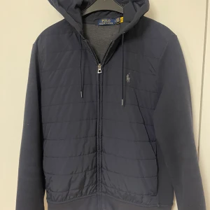 Mörkblå jacka från Polo Ralph Lauren - Snygg mörkblå jacka från Polo Ralph Lauren med huva och dragkedja. Jackan har ett quiltat mönster på framsidan och långa ärmar. Perfekt för kyligare dagar. Den är i storlek M