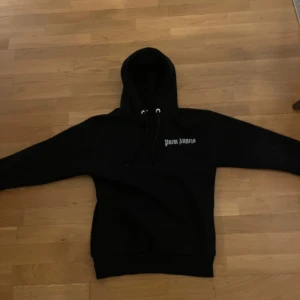 Svart hoodie från Palm Angels - Säljer en svart hoodie från Palm Angels med stor logga på ryggen och mindre logga på bröstet. Tröjan har en klassisk design med huva och dragsko. Perfekt för en avslappnad stil.