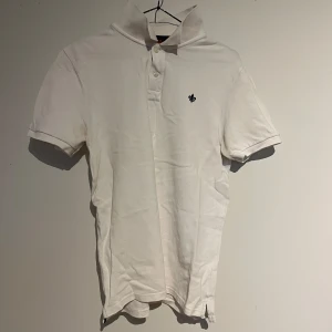 Vit pikétröja från Morris - Snygg vit pikétröja från Morris med korta ärmar och klassisk krage. Tröjan har en liten broderad logotyp på bröstet och knappar framtill. Perfekt för en stilren look.