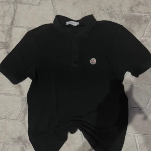 Svart pikétröja från Moncler - Säljer en stilren svart pikétröja från Moncler med korta ärmar och klassisk krage. Tröjan har en broderad Moncler-logga på bröstet och knappar framtill. Perfekt för en avslappnad men ändå elegant look.