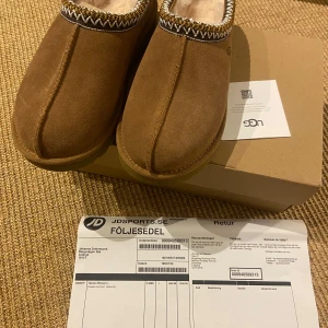 UGG Tasman chestnut  - Julklapp som var för små. Aldrig använda och kvitto finns men retur perioden slut. Säljer ett par bruna UGG tofflor med mjukt foder och dekorativ kant. Perfekta för att hålla fötterna varma och bekväma. De har en robust sula för extra grepp.