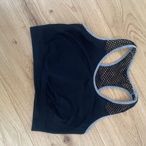 Svart sporttopp från Work Out - Säljer en svart sporttopp från Work Out med grå kantdetaljer och meshpaneler för extra ventilation. Perfekt för träning och ger bra stöd. Toppen har en racerback-design för ökad rörelsefrihet.