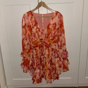 Färgglad kortklänning med volanger (Sanna Jörnvik x NAKD) - Säljer en färgglad kortklänning med volanger i orange och rosa toner. Klänningen har en v-ringad design och långa ärmar. Perfekt för att sticka ut med sitt unika mönster och flödande stil. Från Sanna Jörnviks kollektion med NAKD.