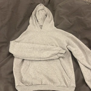 Grå hoodie - Säljer en klassisk grå hoodie med ficka framtill och huva. Perfekt för en avslappnad stil. Den har långa ärmar och ribbade muddar vid ärmslut och nederkant.
