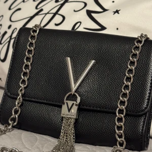 Svart axelväska med kedja från Valentino - Snygg svart axelväska i skinn från Valentino med en stilren kedja och ett stort V på framsidan. Väskan har en praktisk dragkedja inuti och är perfekt för att bära dina viktigaste saker. En elegant accessoar för alla tillfällen.