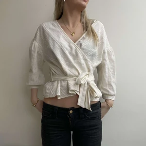 Vit omlottblus med knytband - Säljer en stilren vit omlottblus med knytband i midjan. Blusen har trekvartsärmar och en v-ringad design som ger en elegant touch. Perfekt för både vardag och speciella tillfällen.