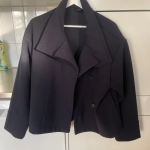 Marinblå kappa - Snygg svart kappa med dubbelknäppning och bred krage. Den har en kortare passform och långa ärmar, perfekt för en stilren look. Passar bra till både vardag och mer uppklädda tillfällen. Den är ganska ny och inga tecken på användning 