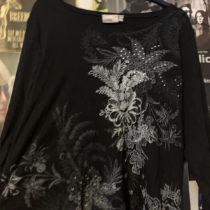 Svart långärmad topp från Isolde - Snygg svart långärmad topp från Isolde med ett vackert blommönster i grått och silverdetaljer. Nästan likt affliction! Perfekt för att ge en elegant touch till din outfit. 🌟