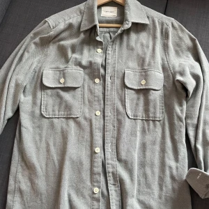 Grå overshirt från A Day's March - Snygg grå overshirt från A Day's March med knappar framtill och två bröstfickor. Perfekt för lager-på-lager-stil. Klassisk krage gör den mångsidig och stilren. Materialet är 100% bomull.