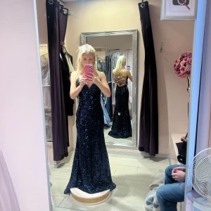 Mörkblå glittrig balklänning  - Säljer en fantastisk mörkblå långklänning med glittriga paljetter. Klänningen har en v-ringad design och en öppen rygg med snörning. Perfekt för en glamorös kväll! Klänningen kan även snöras på flera olika sätt i ryggen. Klänningen är aldrig använd utan bara testad då jag beslutade mig för en annan klänning istället. Jag köpte klänningen för 5000 kr.