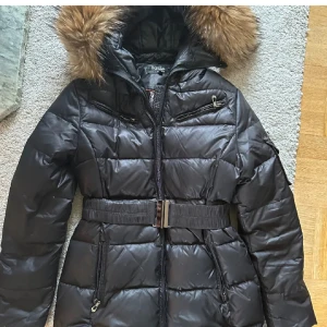 Svart dunjacka från Hollies - Snygg svart dunjacka från Hollies med pälskantad huva och bälte i midjan. Orginalpris ligger på 4000