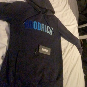 Mörkblå hoodie från Hoodrich - Säljer en snygg mörkblå hoodie från Hoodrich med en stor ficka framtill och justerbar huva. Hoodien har texten 'HOODRICH' i blå nyanser på bröstet. Perfekt för en avslappnad stil.