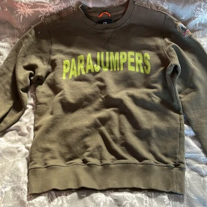 Olivgrön sweatshirt från Parajumpers - Säljer en olivgrön sweatshirt från Parajumpers med stor logga i gult på framsidan. Tröjan har långa ärmar och ribbade muddar vid ärmslut och nederkant. 