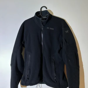 Svart fleecejacka från Arc'teryx - Säljer en svart fleecejacka från Arc'teryx med dragkedja framtill och två praktiska fickor. Jackan har en hög krage och är perfekt för kyligare dagar. Den är stilren och funktionell, idealisk för utomhusaktiviteter.