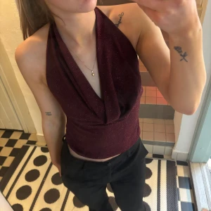 Vinröd glittrig halterneck topp - Snygg vinröd halterneck topp med glittrigt material. Perfekt för en kväll ute eller fest. Toppen har en djup urringning och är figurnära för en smickrande passform.