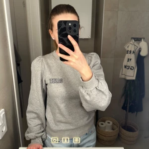 Grå sweatshirt från Ganni - Säljer en stilren grå sweatshirt från Ganni med puffiga ärmar och fina detaljer. Tröjan har en broderad logotyp på bröstet. Säljer då jag tycker den är lite kort på mig, i nyskick. Strl XXS.