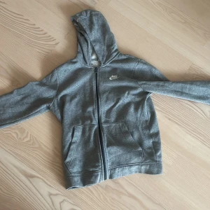 Grå hoodie från Nike - Säljer en grå hoodie från Nike med dragkedja och huva. Den har en klassisk design med två fickor framtill och Nike-loggan på bröstet. Perfekt för en avslappnad stil.