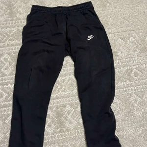 Raka Nike byxor  - Säljer ett par svarta mjukisbyxor från Nike med elastisk midja och det klassiska Nike-loggan i vitt på benet. Perfekta för en avslappnad stil eller träning. Byxorna har en bekväm passform och är gjorda i mjukt material.