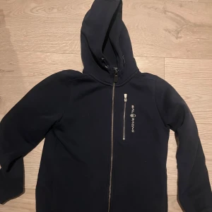 Mörkblå hoodie från Sail Racing 160 - Säljer en mörkblå hoodie från Sail Racing med dragkedja och huva. Perfekt för kyliga dagar. Den har en stilren design med logotyp på bröstet.