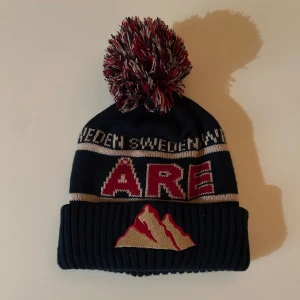 Mössa med tofs och Åre-broderi - Snygg mössa i mörkblått med stor tofs och broderad text 'Åre' i rött. Mössan har även ett bergsmotiv och texten 'Sweden' i vitt. Perfekt för kalla dagar.