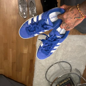 Blå Adidas Campus sneakers - Snygga blå Adidas Campus sneakers med vita ränder och snörning. Perfekta för en casual look med sin klassiska design och bekväma passform.
