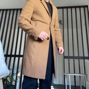 Beige kappa från Zara - Säljer en stilren beige kappa från Zara i ull. Kappan har en klassisk design med knappar framtill och långa ärmar. Perfekt för höst och vinter. Tillverkad med hållbart tyg från Italien.