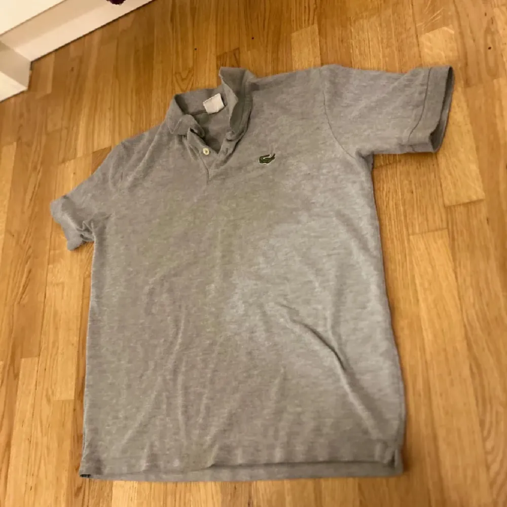 Snygg grå pikétröja från Lacoste med klassisk krage och knappslå. Perfekt för en avslappnad stil. Den har korta ärmar och den ikoniska krokodilloggan på bröstet.. T-paidat.