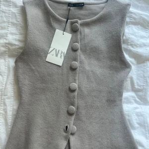Beige väst från Zara - Säljer en stilren beige väst från Zara med knappar framtill. Västen har en klassisk design och är perfekt för lager-på-lager-stil. Den är tillverkad i ett mjukt material som ger en bekväm passform. Helt ny med prislapp kvar