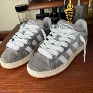 Grå Adidas campus 00s - Snygga grå Adidas sneakers i mocka med vita detaljer och snörning. Skorna har en klassisk design med den ikoniska tre-randiga looken på sidorna. Perfekta för en stilren vardagslook.