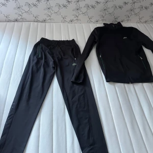 Svart set från Lacoste - Snyggt svart set från Lacoste! På en av byxfickorna har taget på dragkedjan åkt av men funkar ändå att dra uppochner. Köpt från Lacoste och säljs då det inte riktigt passar min stil! Pris kan diskuteras!