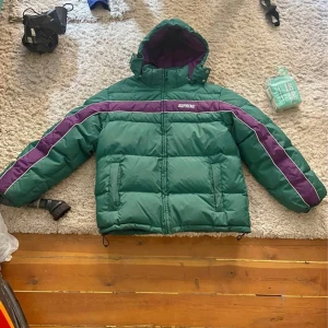 Grön och lila dunjacka från Supreme - Säljer en cool grön dunjacka från Supreme med lila detaljer. Jackan har en dragkedja framtill och en huva för extra värme. Perfekt för kalla dagar när du vill hålla stilen. 💚💜