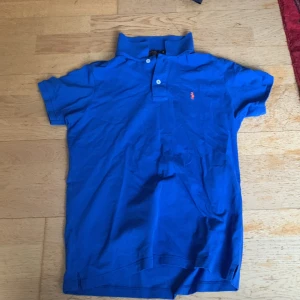 Blå pikétröja från Ralph Lauren - Säljer en klassisk blå pikétröja från Ralph Lauren med den ikoniska broderade loggan i orange på bröstet. Tröjan har korta ärmar och en stilren krage med knappar. Perfekt för en avslappnad och stilren look. Den passar S och XS