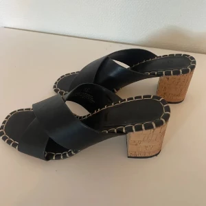 Svarta sandaler med korkklack - Snygga svarta sandaler med korsade remmar och en unik korkklack. Sandalerna har dekorativa sömmar längs kanterna som ger en extra touch. Perfekta för en stilren look.