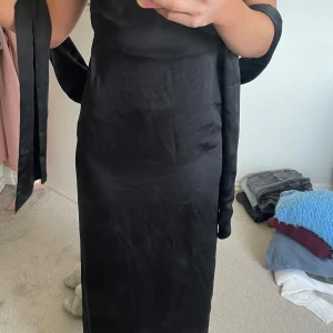 Svart långklänning med öppen rygg - Elegant svart långklänning i satin med öppen rygg och smala axelband. Klänningen har en stilren design med knytband i midjan för en smickrande passform. Perfekt för en festlig kväll. Banden i midjan går att ha lite hur man vill. Klänningen går hela vägen ner till marken och lite lång för mig som är 170. Aldrig använd 